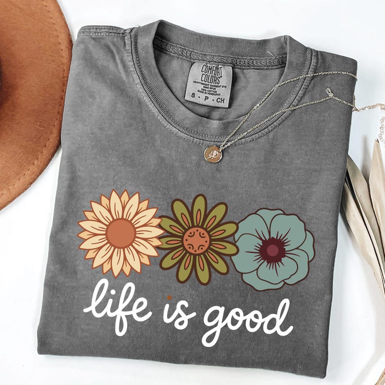 May include: A gray t-shirt with a floral design and the phrase "life is good" in white script. The shirt features three flowers in shades of orange, green, and blue. A silver necklace with a small pendant is draped across the shirt.