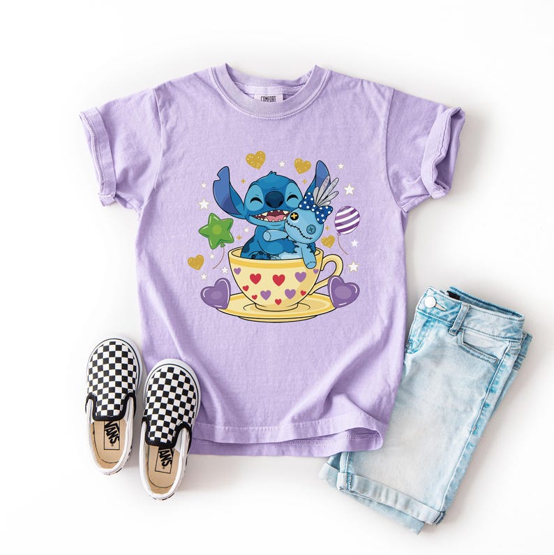 May include: A lilac t-shirt with a cartoon illustration of Stitch and a doll in a teacup, surrounded by hearts, stars, and a balloon. The image is accompanied by checkered slip-on shoes and denim shorts.