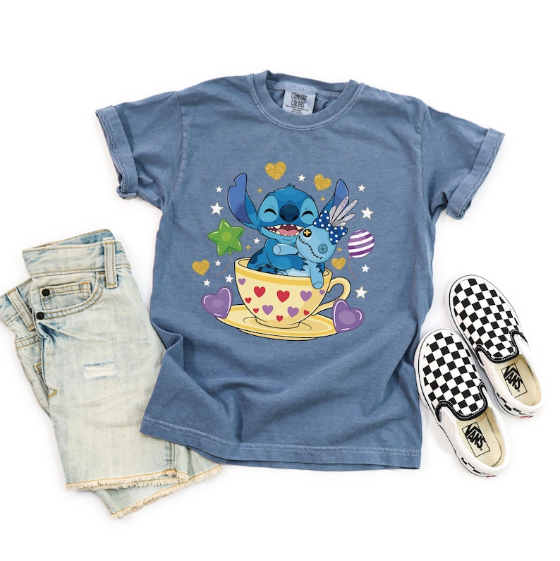 May include: A blue t-shirt with a cartoon Stitch design in a teacup. The teacup is yellow with red hearts. The shirt is styled with denim shorts and black and white checkered slip-on shoes.