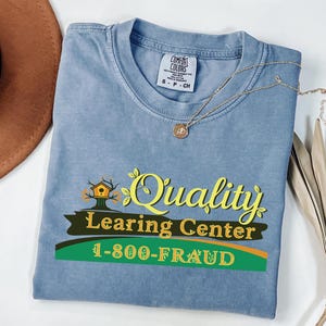 May include: A blue Comfort Colors t-shirt with the words "Quality Learning Center" and the phone number "1-800-FRAUD" printed on the front. The shirt also features a birdhouse graphic and a gold necklace.