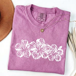 May include: A mauve Comfort Colors t-shirt with a white floral design of three hibiscus flowers. The shirt has a crew neck and a small tag at the neckline. A gold necklace with a pendant rests on the shirt.