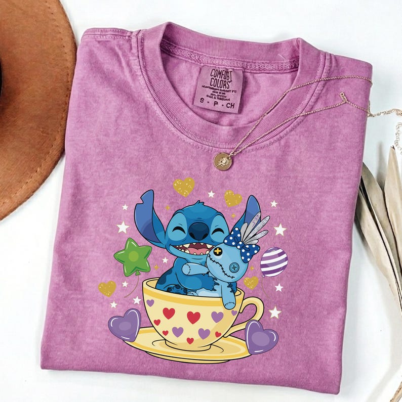 May include: A purple Comfort Colors t-shirt with a graphic of Stitch from Lilo & Stitch. Stitch is in a teacup with a doll. The design includes hearts, stars, and balloons. A gold necklace is draped over the shirt.