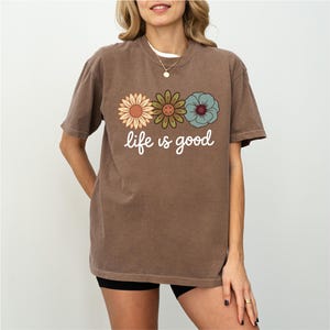 May include: Brown t-shirt with the phrase "life is good" in white script. The shirt features three colorful flower graphics: a sunflower, a green flower, and a blue flower. The shirt is a casual style.