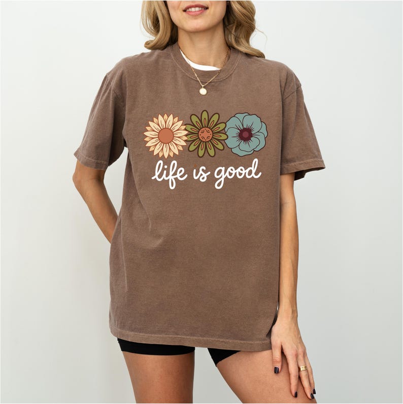 May include: Brown t-shirt with the phrase "life is good" in white script. The shirt features three colorful flower graphics: a sunflower, a green flower, and a blue flower. The shirt is a casual style.