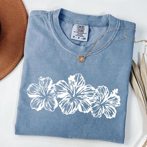 May include: A blue Comfort Colors t-shirt with a white floral design. The shirt has a crew neck and short sleeves. A gold necklace with a pendant is draped across the shirt. The shirt is size S-P-CH.