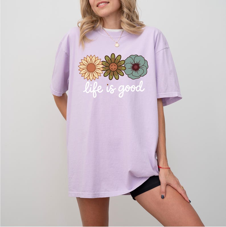 May include: A light purple t-shirt with three retro-style flowers in yellow, green, and teal. The words "life is good" are printed in white script below the flowers. The shirt is a casual, oversized fit.