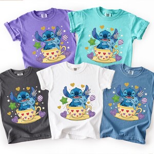 May include: Five short-sleeved t-shirts in purple, teal, white, charcoal grey, and blue. Each shirt features a cartoon illustration of a blue alien character in a teacup, surrounded by hearts and stars.