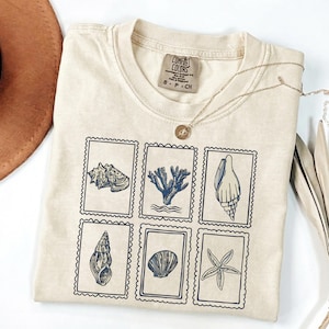 May include: Cream-colored t-shirt featuring six postage stamp-style illustrations of seashells and coral in navy blue. The shirt has a crew neck and short sleeves. A gold necklace with a pendant is draped across the shirt.