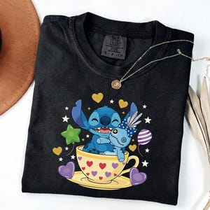 May include: Black Comfort Colors t-shirt with a Stitch cartoon design in a teacup, holding a doll. The design includes hearts, stars, and balloons. A gold necklace is draped across the shirt.