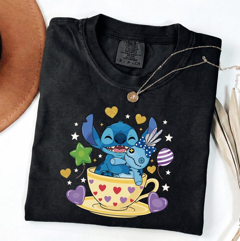 May include: Black Comfort Colors t-shirt with a Stitch cartoon design in a teacup, holding a doll. The design includes hearts, stars, and balloons. A gold necklace is draped across the shirt.