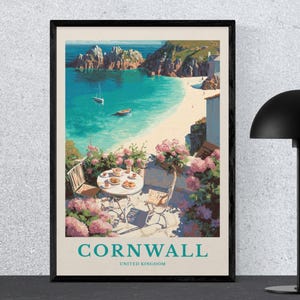 May include: A framed art print of Cornwall, United Kingdom. The print depicts a coastal scene with turquoise water, a sandy beach, and rocky cliffs. A table set for two is on a terrace, surrounded by pink flowers. The print has a black frame.