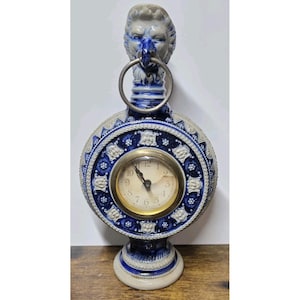 May include: A ceramic clock with a blue and white floral design. The clock has a round face with gold-colored trim and a lion's head at the top. The clock is approximately 10 inches tall.