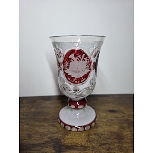 May include: A clear and ruby red glass goblet featuring cut glass details and a central oval medallion. The medallion displays an etched illustration of an open book and musical instruments with the text u. Freude inscribed below the design.
