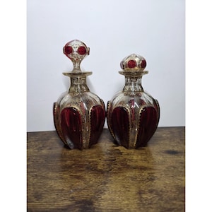May include: Two matching glass decanters featuring deep red panels and clear glass sections with intricate gold filigree detailing. Each decanter has a rounded body, a narrow neck, and a matching stopper with red glass accents and gold trim.