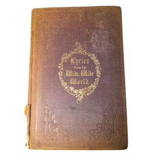 May include: A brown hardcover book with gold embossed lettering on the front cover that reads Lyrics from the Wide, Wide World. The cover features a central floral wreath design and decorative corner scrollwork patterns on the front board.