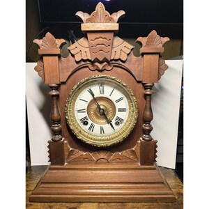 May include: A brown wooden mantel clock featuring a circular face with black Roman numerals, a gold-toned decorative bezel, and two black hands. The clock case has carved architectural details, including finials and a stepped base design.
