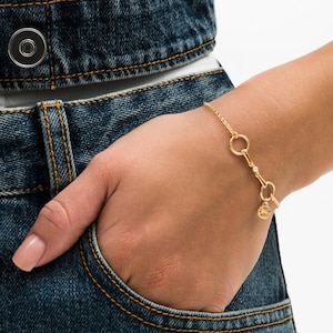 May include: A gold-toned chain bracelet featuring a central bar element with a small embedded clear stone, two circular links, and a round dangling charm with a textured surface, secured with a delicate link chain around the wrist.