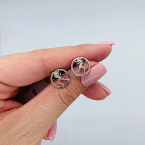 May include: A pair of silver-toned stud earrings, each featuring a circular frame enclosing a detailed silhouette of a figure in a dynamic running pose. The earrings have a polished metallic finish and are designed with a post-back closure mechanism.
