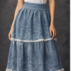 May include: A woman wearing a white top with ruffled sleeves and a long, denim blue skirt. The skirt has a tiered design with a frayed hem and a white, swirling pattern. The outfit is casual and stylish.