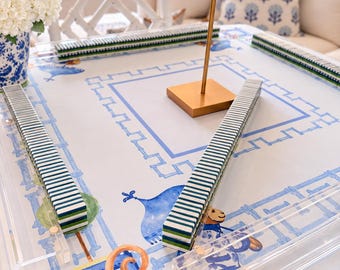 May include: A square mahjong table cover featuring a white background with blue bamboo-style border patterns and whimsical blue and orange illustrations. Four clear acrylic tile racks are positioned on the surface, each holding a row of green and white tiles.