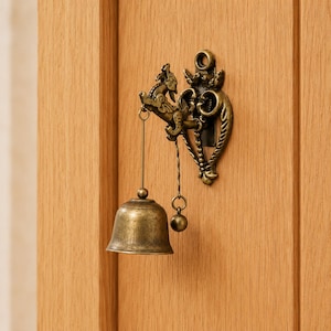 May include: Vintage brass doorbell with an intricate design of a mythical beast and decorative flourishes. The bell hangs from a chain and is mounted on a wooden door. A classic door accessory.