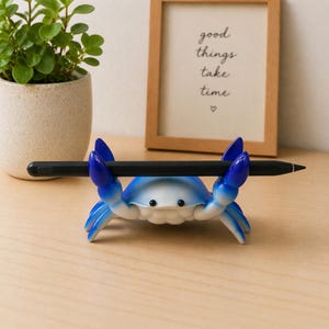 May include: A blue and white crab-shaped pen holder with a black pen. The crab has blue claws and a smiling face. A framed picture with the text "good things take time" is in the background.