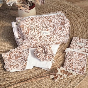 May include: A quilted toiletry bag, a matching scrunchie, a set of square facial pads, and a rectangular fabric pouch, all featuring a brown floral print on a white background. Each item includes a small white label with black text.