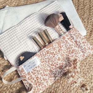 Peut inclure: Un sac de maquillage à imprimé floral avec une poignée, contenant des pinceaux de maquillage, des rouges à lèvres et un compact. Le sac est ouvert, révélant le contenu, et est posé sur une serviette blanche gaufrée. Le texte "Un Fil de Luciole Fait main" est visible.