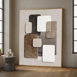 May include: Abstract wall art featuring a gold frame and a neutral color palette. The artwork showcases layered, textured squares in shades of brown, beige, and white, creating a modern and minimalist design. The piece is displayed in a well-lit room.