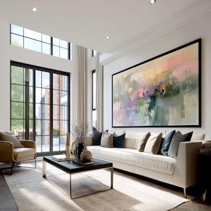 May include: A modern living room featuring a large abstract painting with a black frame. The painting has soft pastel colors. A white sofa with decorative pillows sits below the artwork. A black coffee table and a beige armchair complete the room.