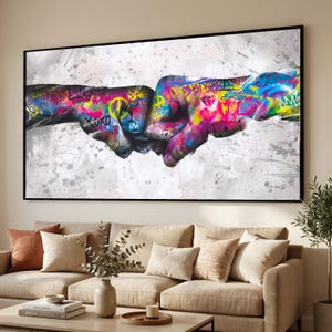 May include: A large framed canvas art piece featuring two fists bumping, painted with vibrant graffiti art. The artwork uses a palette of bright colors including pink, yellow, blue, and green, set against a white and gray splattered background. The piece is displayed above a beige sofa.