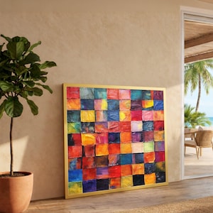 May include: A large, framed artwork with a colourful patchwork design. The artwork is made up of many squares in shades of red, orange, yellow, blue, and purple. The frame is gold, and the artwork leans against a neutral wall.