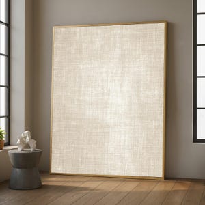 May include: Large rectangular canvas art with a gold frame. The canvas has a neutral, off-white linen texture. The artwork is leaning against a wall in a room with hardwood floors and a small side table.