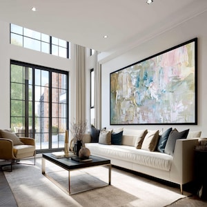 May include: A modern living room featuring a large abstract painting with a black frame. The painting has a textured appearance with pastel colors. A white sofa with decorative pillows sits below the artwork. A black coffee table and a tan armchair complete the space.