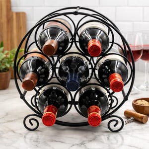 May include: A black metal wine rack holding several bottles of wine. The rack has a circular design with curved accents. The wine bottles are visible, with red and brown bottle tops. A corkscrew and wine glasses are in the background.