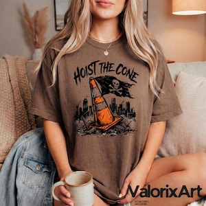 May include: Brown t-shirt with the text "HOIST THE CONE" above a graphic of an orange traffic cone with a skull and crossbones flag. The graphic includes a cityscape and rubble.