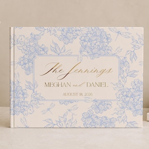May include: A rectangular hardcover book with a cream-colored cover featuring a light blue floral and dragonfly pattern. The center has a rectangular frame containing the gold text: The Jennings, MEGHAN and DANIEL, AUGUST 18, 2026.