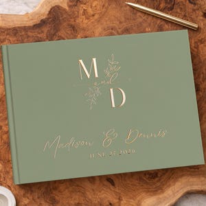 May include: A sage green hardcover guest book featuring gold foil lettering that reads M and D with a botanical illustration, followed by the names Madison & Dennis and the date JUNE 27 2026 in a script and sans-serif font combination.
