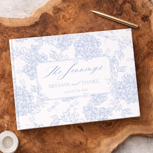 Op de afbeelding: Een wit gastenboek met een blauw bloemmotief en de tekst "The Jennings". Het toont de namen "Meghan en Daniel" en de datum "18 augustus 2026". Een gouden pen ligt op het houten oppervlak naast het boek.
