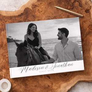 以下が含まれることがあります： 「Madison & Jonathan」の名前がエレガントな筆記体で書かれた白黒のゲストブック。表紙には、女性が馬に乗っているビーチでのカップルの写真が写っています。金のペンが木の表面に置かれています。
