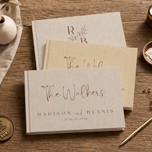 May include: Three linen-covered wedding guest books with custom text, stacked on a wooden surface. The top book has the initials "R" and "B" with a floral design. The bottom book reads "The Walkers, Madison and Dennis, June 27, 2026."