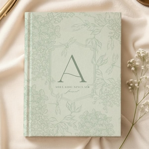 Puede incluir: Un diario de tapa dura con un diseño floral verde claro y la letra "A" en un escudo. El diario tiene el texto "Adelaide Sinclair Journal" y está rodeado por un patrón floral. Un bolígrafo dorado y flores blancas están en el fondo.