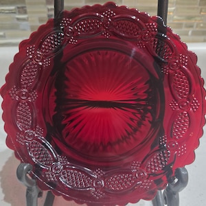May include: A vibrant ruby red glass plate with a scalloped edge and detailed embossed patterns. The plate has a central sunburst design and decorative oval motifs around the rim. It is displayed on a dark metal stand, highlighting its vintage appeal.