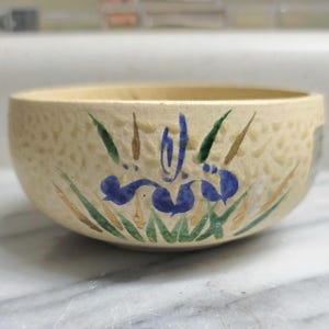 Puede incluir: Un cuenco de cerámica de color crema con una superficie exterior texturizada y con hoyuelos. La parte frontal presenta un diseño pintado a mano de una flor de iris azul con hojas verdes y tallos marrones. El cuenco tiene una abertura ancha.