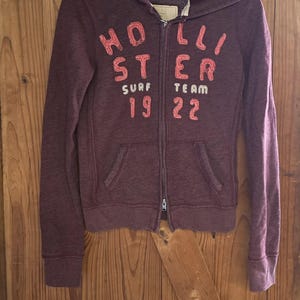 May include: A dark maroon zip-up hoodie featuring a hood, long sleeves, and two front pockets. The front displays the text HOLLISTER SURF TEAM 19 22 in a textured, light red font. The garment is displayed on a black plastic hanger.