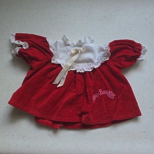 May include: A red velvet doll top featuring a white eyelet lace collar with a cream ribbon tie at the centre. The sleeves are short and puffed with white lace trim. The text Cabbage Patch Kids is embroidered in pink on the lower right side.