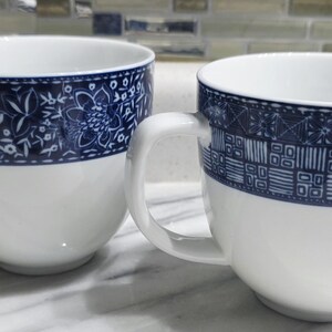 Könnte beinhalten: Zwei weiße Keramikbecher mit einem blauen floralen und geometrischen Musterband am oberen Rand. Jeder Becher hat einen großen, quadratischen Griff. Die Becher stehen auf einer weißen Oberfläche.