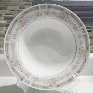 May include: A white ceramic bowl with a floral pattern. The rim features a gray border with delicate pink, yellow, and purple flowers. The bowl is displayed on a clear plastic stand, showcasing its elegant design.
