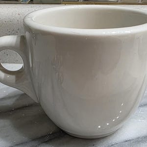 May include: A white ceramic coffee cup with a rounded body and a large handle. The cup has a smooth, glossy finish and is designed for holding hot beverages. The cup is empty and ready to be filled.