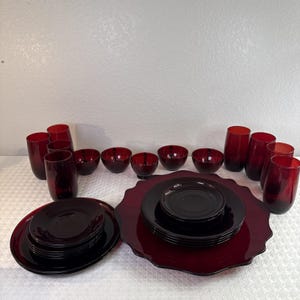 May include: A collection of deep red glassware including several tall drinking glasses, small bowls, and two stacks of plates. One stack features smooth round plates, while the other rests on a large, scalloped-edge red glass serving platter.